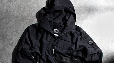 ST95 JP-8 Anorak in black, featuring a hooded design, front zip pockets, and the signature sleeve logo badge.
