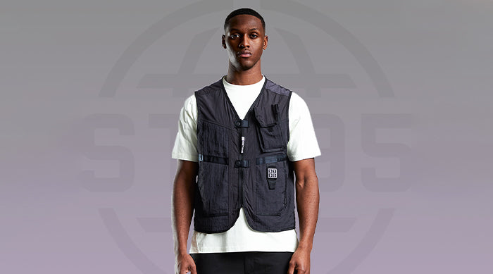 Model wearing the ST95 Delta Vest in black with utility pockets and front zip, styled over a white T-shirt against grey backdrop.