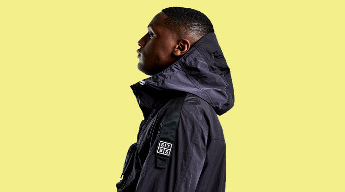Side profile of model wearing the ST95 Drogue Smock 2.0 in black, featuring hood and sleeve logo patch, against yellow background.
