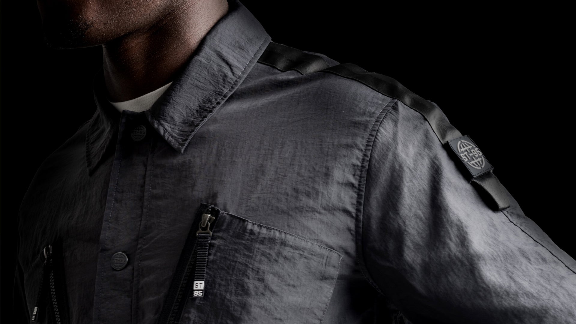 Close-up of the ST95 JP-8 Overshirt in dark grey, showing chest zip pocket, button detail, and branded sleeve badge.